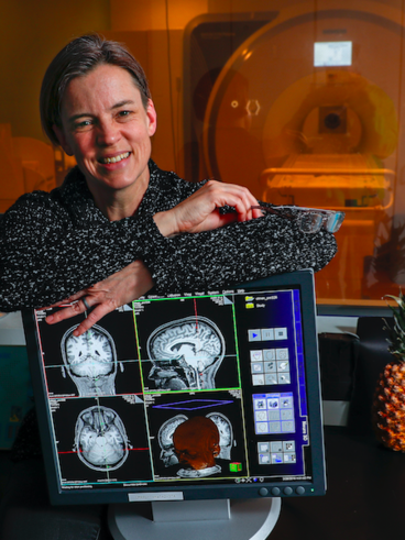 Cheryl sitting in front of a 3T magnet holding a console monitor showing a T1-weighted image of her brain.
